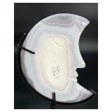 A luminous Botswana Agate druzy carved into a moon shape, showcasing delicate bands of soft gray, caramel, and cream. The sparkling druzy surface catches the light, in a custom metal stand.