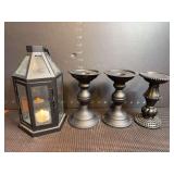 Three black metal pedestal-style pillar candle holders and one black metal and glass lantern-style candle holder with a battery operated tea light candle