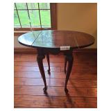 Oval drop-leaf table with cherry finish and turned legs. Features a compact design with hinged leaves that can be raised for additional surface space or lowered for storage. Traditional accent or occa