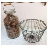 Vintage wire egg basket accompanied by a large glass jar with wire-and-wood carrying handle filled with pine cones.