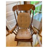 Wood rocking chair with spindle back, shaped arms and brown tufted seat and back cushions. Shows age-related surface wear.