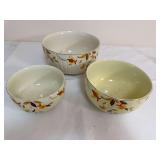 Three Hall Autumn Leaf mixing bowls in graduated sizes with gold trim and the familiar floral decoration. The bowls show noticeable differences in glaze/color, ranging from a lighter ivory to a more y