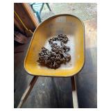 Vintage metal wheelbarrow with yellow/orange painted tray and wood handles, containing a length of heavy chain with hooks as shown. Shows paint wear, oxidation and age-related use.