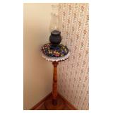 Turned wood pedestal plant stand with round top, accompanied by a vintage oil lamp with decorative dark metal base and clear glass chimney, plus floral decoration as shown.