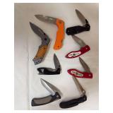 Eight folding pocket and utility knives including NRA/NRA-ILA promotional examples and two red firefighter-themed knives. Includes plain and partially serrated blades with assorted handle styles.