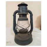 Dietz Little Wizard tubular kerosene lantern with dark painted metal body, clear glass globe and wire bail handle. Shows age-related surface wear, soiling and oxidation.