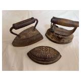 Three antique cast-iron sad irons as shown, including examples with wood handles and one marked “Asbestos Sad Iron.” Shows oxidation and age-related surface wear.