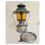 Coleman Model 228D “Big Hat” pressure lantern with silver-tone fount, green porcelain ventilator, amber Coleman Pyrex globe and wire bail frame. Includes round metal accessory/storage plate as sho