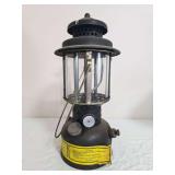 Coleman U.S. Army SMP 1984 pressure lantern with dark military-style fount, black ventilator, clear globe, wire frame and yellow instruction label. Shows paint wear, oxidation and general age-related 