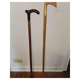 Two walking canes. One is metal with a wood handle 32.5 in tall. The other has a carved shaft and is 35.5 in tall.