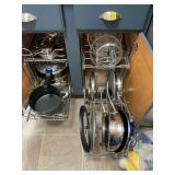 Assortment of nice pots and pans with lids and pressure cooker in cabinet beside refrigerator. Includes Calphalon, Cuisinart, Kirkland and others.