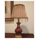 Table lamp, ornate, textured glass font in deep burgundy and red, stepped, antique bronze-finished resin base, topped with a pleated rose fabric empire shade, embellished with shaggy, raffia-like frin