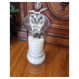 Glass footed apothecary jar with a matching lid, filled with sand, stones, and a decorative butterfly charm hanging from the neck. 16 in tall.