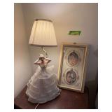 Approx 24” tall porcelain lady table lamp and framed ladies Appears the arm of the lamp has been repaired Holds 2 bulbs so lower half lights up her dress